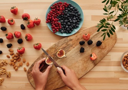 Overhead shot of fresh fruits, nuts, and hands preparing ingredients on a wooden board.